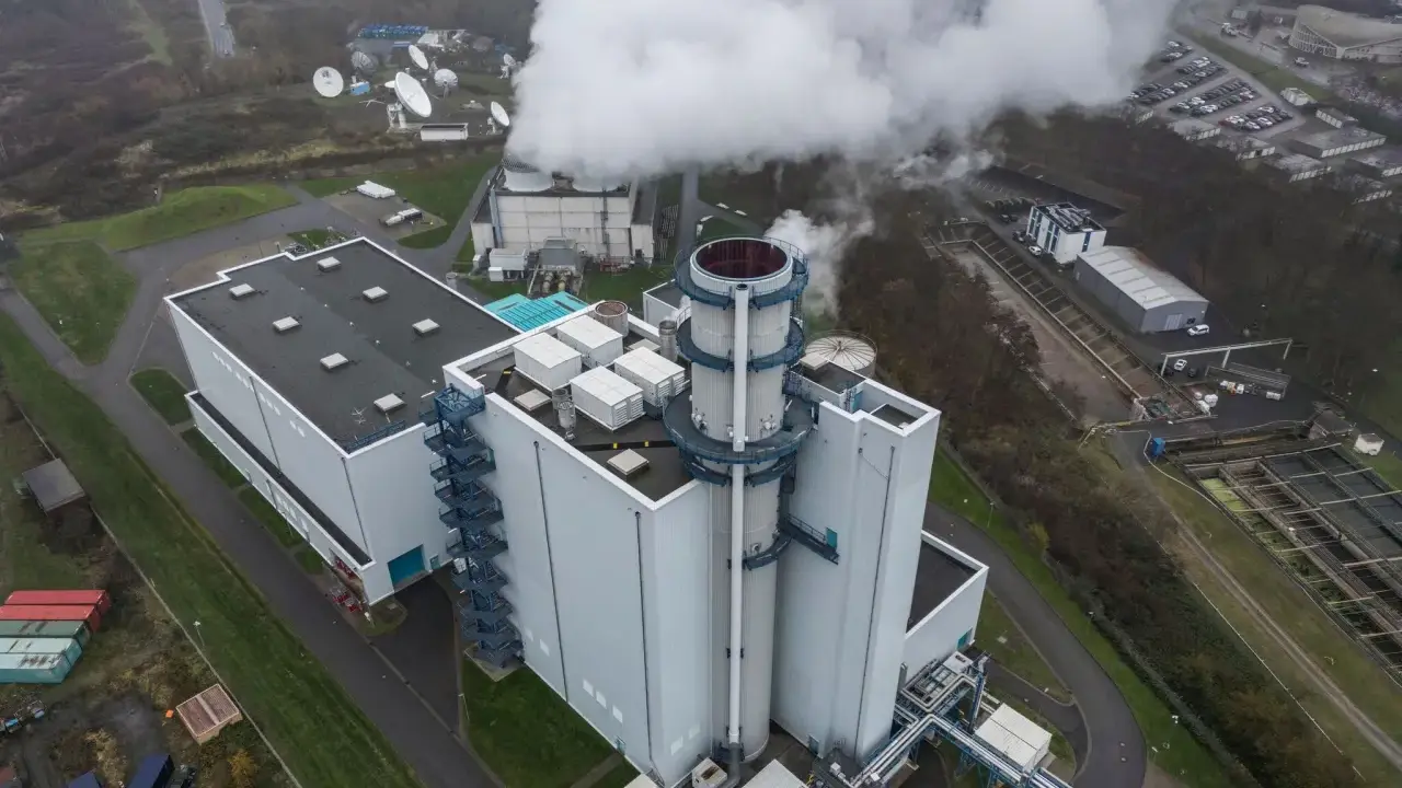 Dampf steigt aus einem modernen Gaskraftwerk in Deutschland auf. Satellitenschüsseln und ein Parkplatz sind in der Nähe zu sehen.