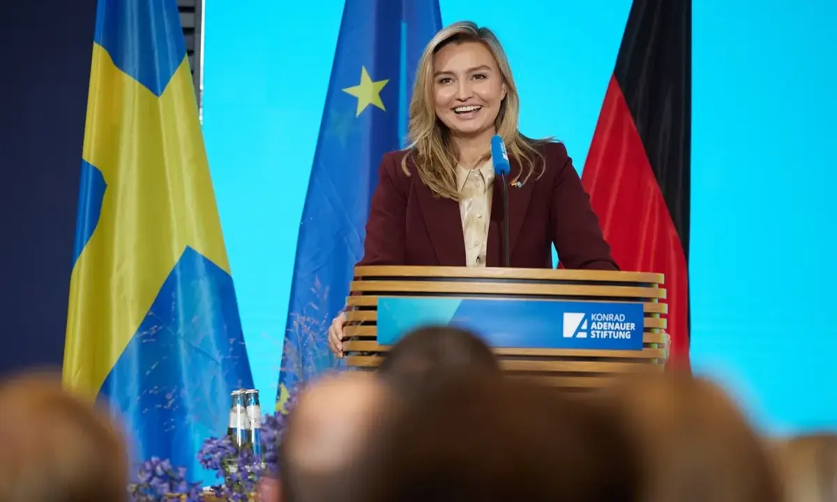 Frau spricht über den Strompreis in Schweden, flankiert von schwedischer und deutscher Flagge.