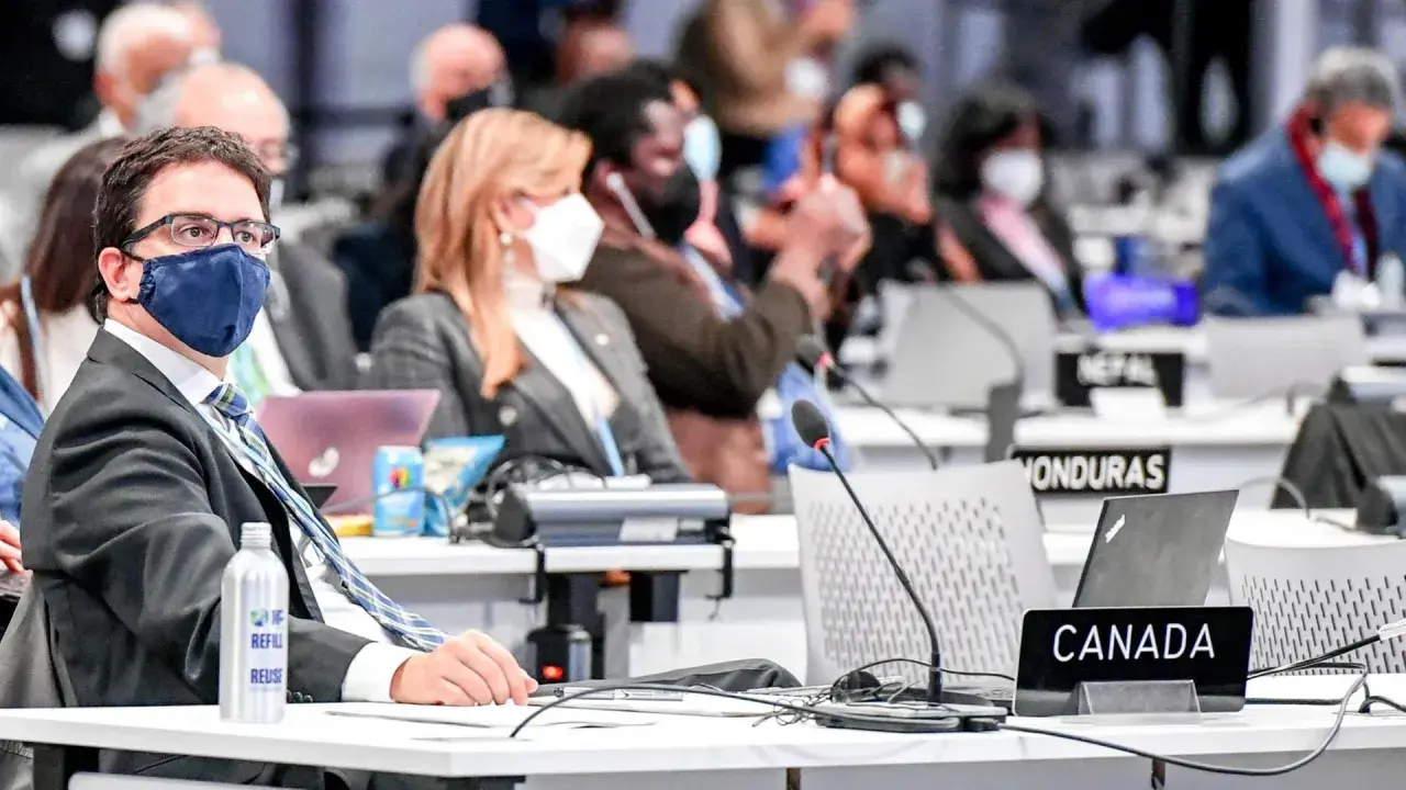 Teilnehmer der Klimakonferenz, darunter ein Mann mit Brille und Maske, sitzen an Tischen mit Laptops und Namensschildern wie "CANADA".