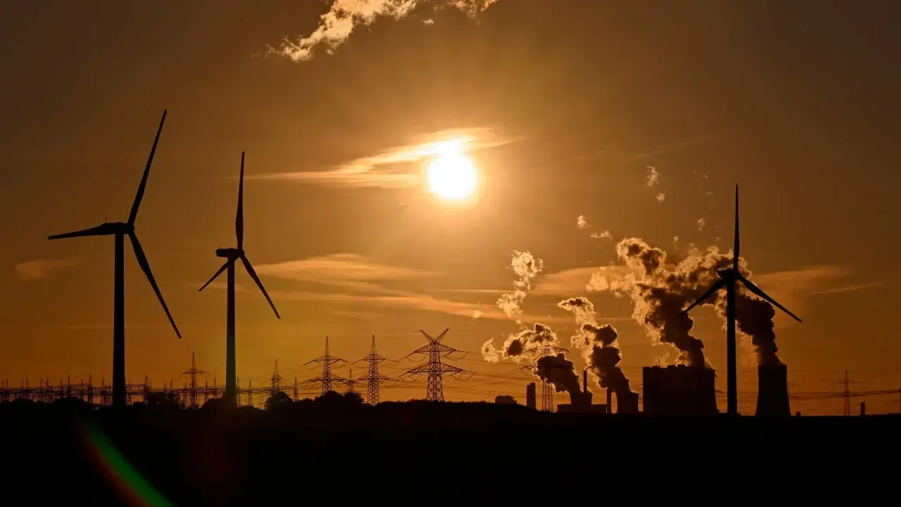 Silhouetten von Windrädern und rauchenden Industrieanlagen im Sonnenuntergang. Ein Bild, das zum Nachdenken über Stromkosten Norwegen vs. Deutschland anregt.