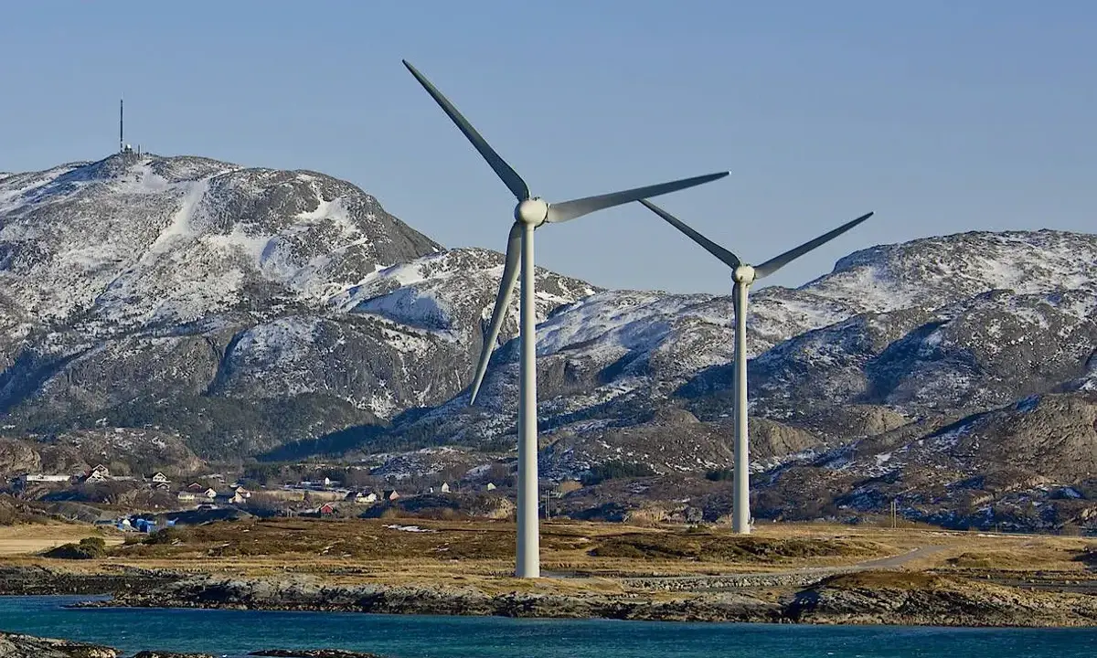 Zwei Windräder erzeugen sauberen Strom aus Norwegen. Im Hintergrund schneebedeckte Berge und ein kleines Dorf.