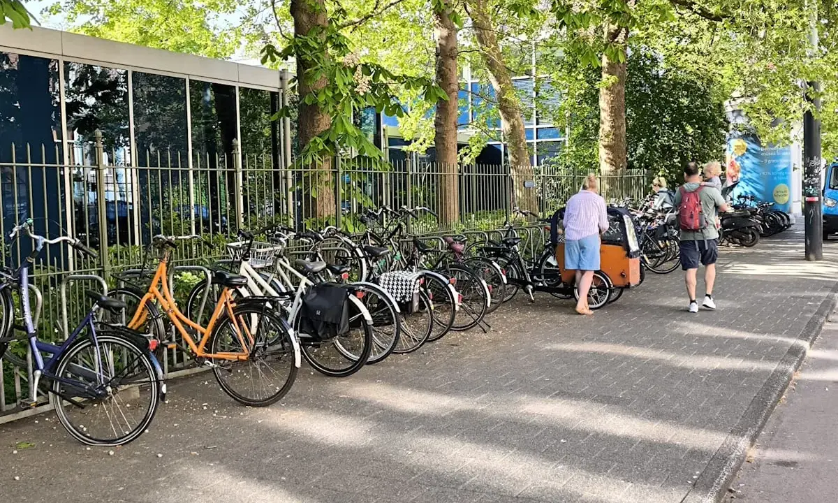 Viele Fahrräder stehen am Straßenrand, ein Mann mit Kind auf dem Arm und eine Frau mit Lastenrad gehen vorbei. Eine echte Stadt der kurzen Wege.