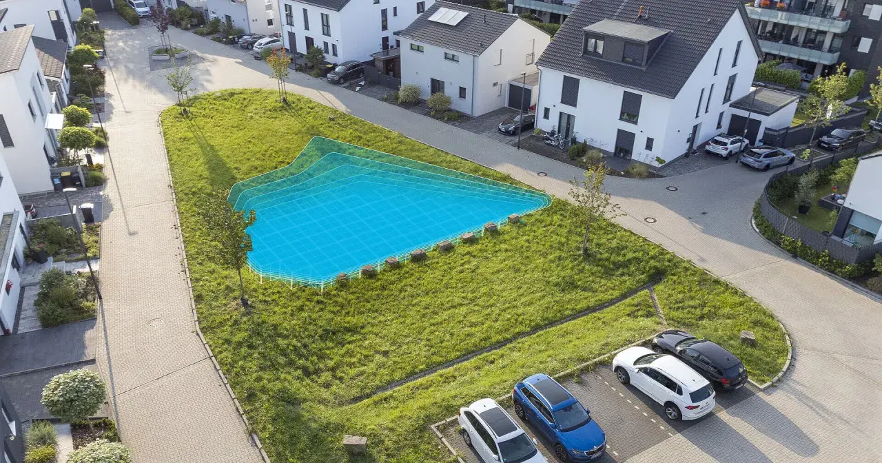 Eine blaue Fläche markiert eine geplante Mulde/Rigole in einem grünen Hang neben modernen Häusern und geparkten Autos.