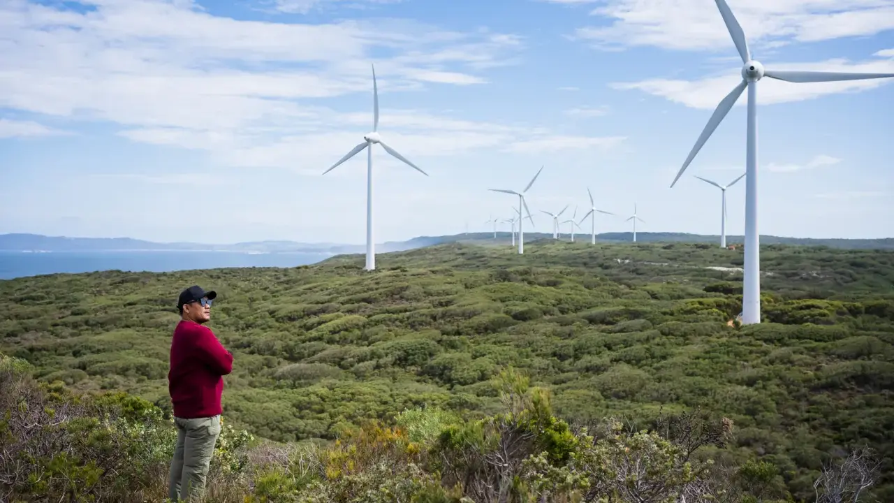 Mann steht vor Windrädern, die die australien energieversorgung repräsentieren. Die Landschaft ist grün und hügelig, mit dem Meer im Hintergrund.