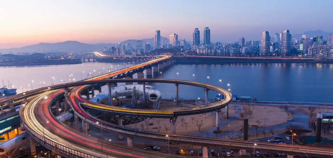 Verkehrsfluss in Seoul bei Nacht, mit Lichtern, die sich auf den Autobahnauffahrten und der Skyline der Stadt spiegeln.