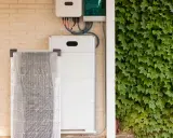 Ein weißer Redox-Flow-Heimspeicher steht neben einer verpackten Solaranlage an einer Ziegelwand, bedeckt mit Efeu.