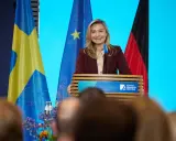 Frau spricht über den Strompreis in Schweden, flankiert von schwedischer und deutscher Flagge.