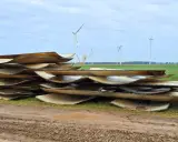 Stapel von Rotorblättern von Windkraftanlagen, bereit für das Recycling. Im Hintergrund sind weitere Windräder zu sehen.