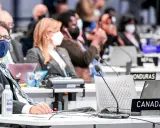 Teilnehmer der Klimakonferenz, darunter ein Mann mit Brille und Maske, sitzen an Tischen mit Laptops und Namensschildern wie "CANADA".