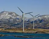 Zwei Windräder erzeugen sauberen Strom aus Norwegen. Im Hintergrund schneebedeckte Berge und ein kleines Dorf.