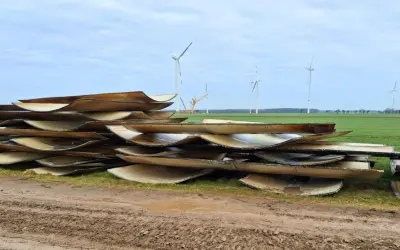 Stapel von Rotorblättern von Windkraftanlagen, bereit für das Recycling. Im Hintergrund sind weitere Windräder zu sehen.
