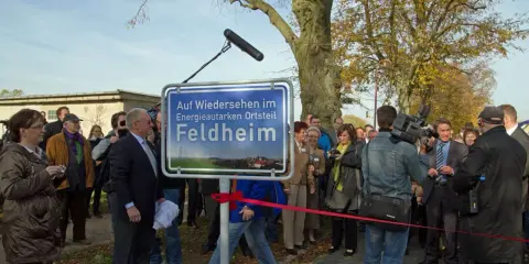 Feldheim, ein Vorreiter unter den energieautarken Gemeinden Deutschlands, wird hier mit einem Schild gefeiert. Eine Gruppe von Menschen, darunter ein Kameramann, versammelt sich.