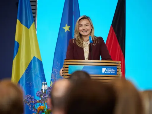 Frau spricht über den Strompreis in Schweden, flankiert von schwedischer und deutscher Flagge.