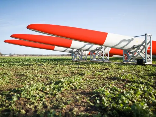 Drei Rotorblätter für Windkraftanlagen liegen auf einem Feld. Sie sind rot-weiß gestreift und warten auf ihren Einsatz nach dem EEG.