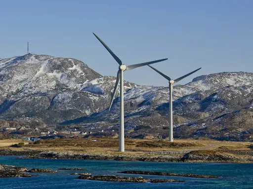 Zwei Windräder erzeugen sauberen Strom aus Norwegen. Im Hintergrund schneebedeckte Berge und ein kleines Dorf.