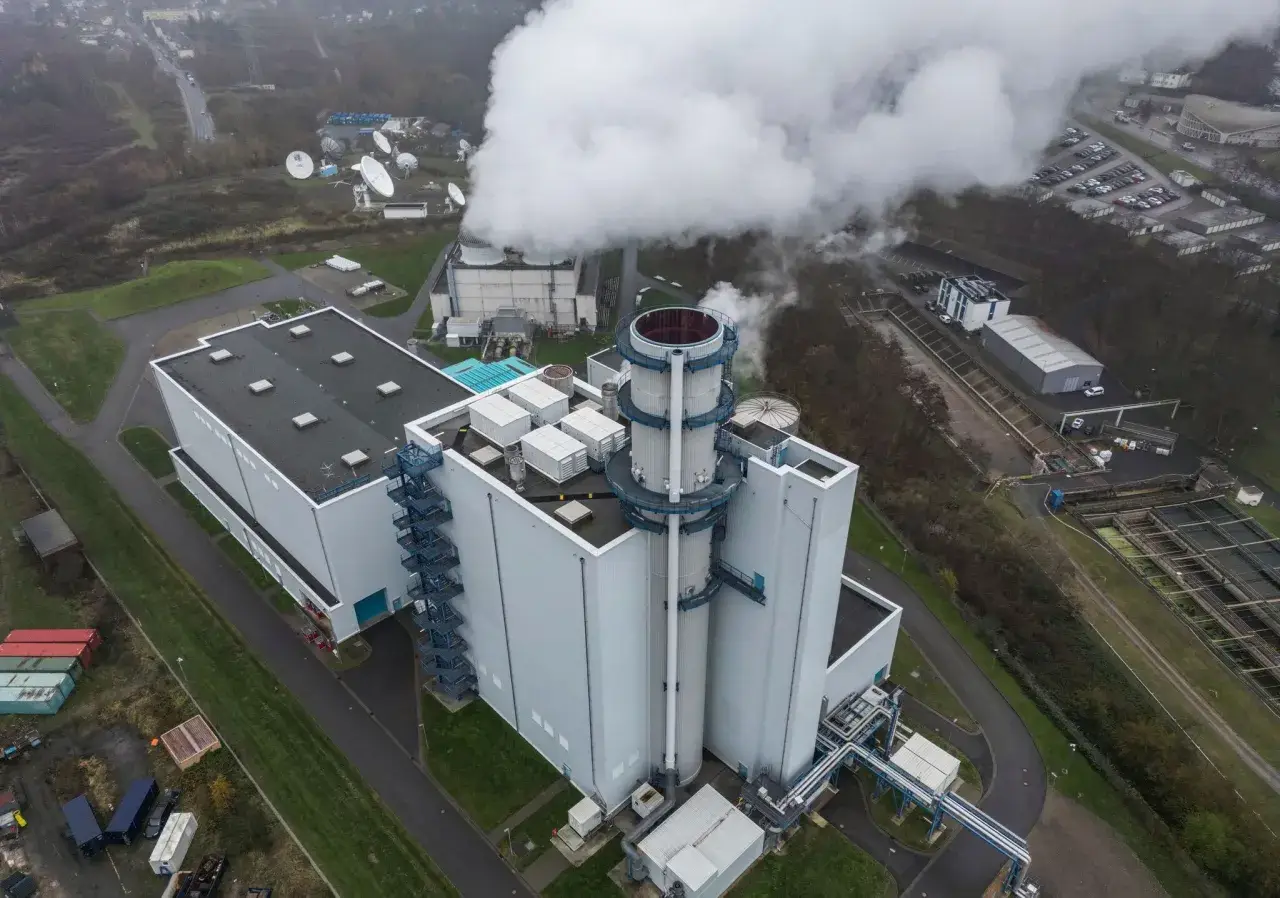 Dampf steigt aus einem modernen Gaskraftwerk in Deutschland auf. Satellitenschüsseln und ein Parkplatz sind in der Nähe zu sehen.