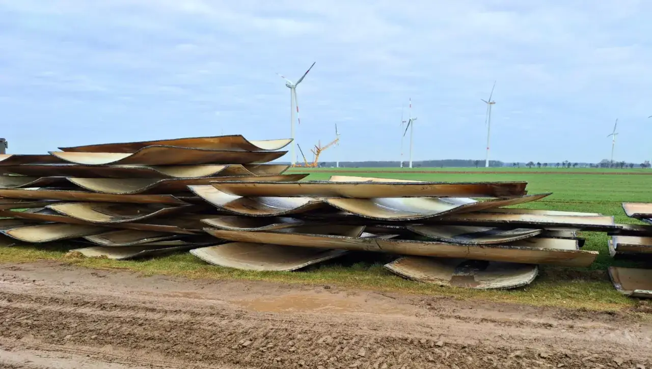 Stapel von Rotorblättern von Windkraftanlagen, bereit für das Recycling. Im Hintergrund sind weitere Windräder zu sehen.