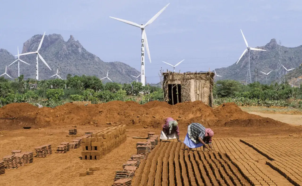 Zwei Frauen stellen Ziegel her, w&auml;hrend im Hintergrund Windr&auml;der f&uuml;r den Klimaschutz in Indien stehen.
