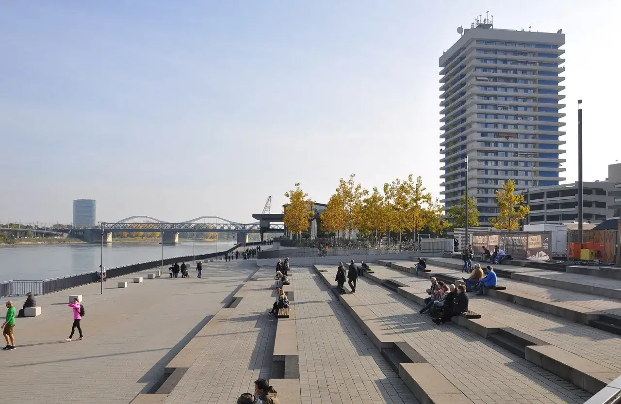 Menschen genießen die Sonne an einem Flussufer mit Brücken und einem Hochhaus. Die weitläufige, gepflasterte Fläche zeigt, was ist versiegelung: eine urbane Gestaltung, die Raum für Erholung schafft.