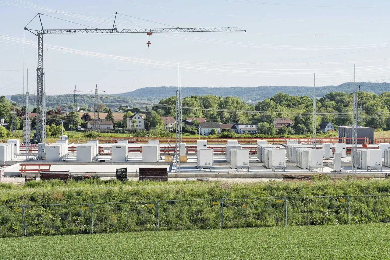 Bau eines großen Stromspeichers in Deutschland. Viele weiße Container stehen auf einer Betonplatte, ein Kran ragt in den Himmel.