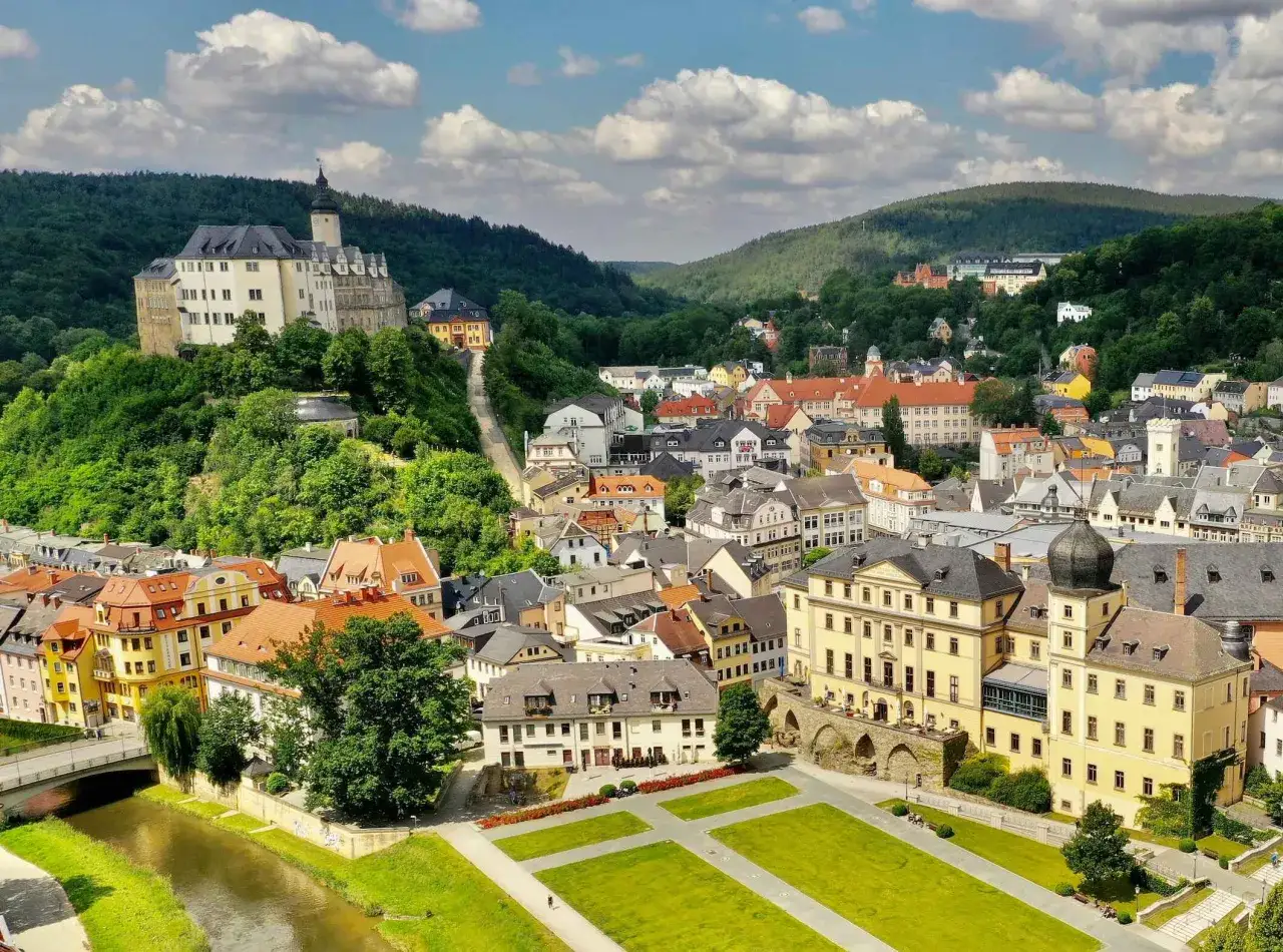 Historische Stadtansicht mit Schloss auf einem Hügel, umgeben von grünen Wäldern und einer malerischen urbanen Entwicklung.