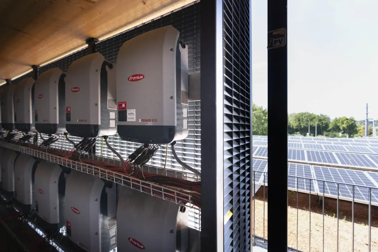 Reihen von Fronius Wechselrichtern, die Teil eines Redox-Flow-Heimspeichersystems sind, sind in einem Gebäude montiert. Dahinter erstrecken sich Solarpanels.