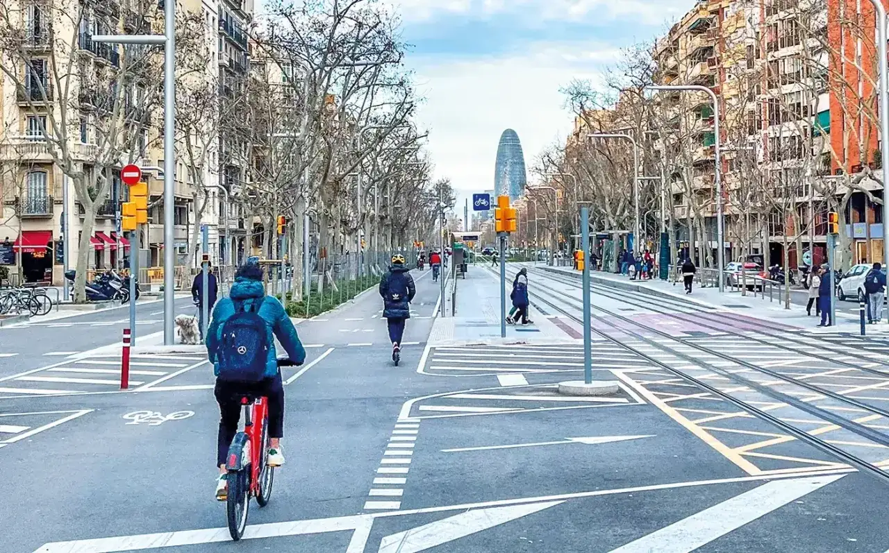Radfahrer und E-Scooter-Fahrer nutzen die breiten Wege in den Superblocks Barcelona. Tramschienen und Bäume säumen die Straße.