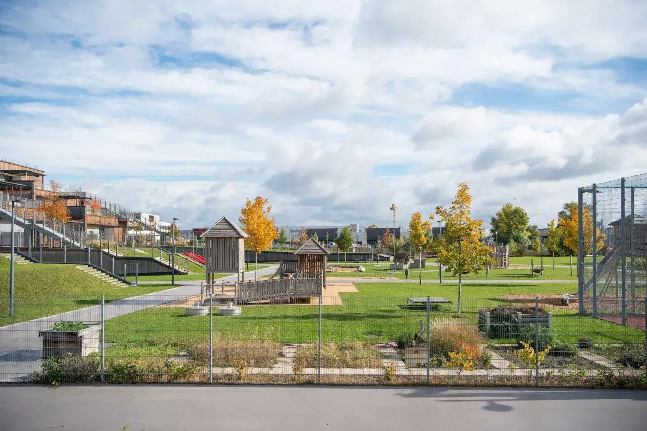 Spielplatz mit Holzspielhäusern, Rutschen und Grünflächen. Moderne Gebäude im Hintergrund. Ein Beispiel für gendergerechte Stadtplanung, die allen zugutekommt.