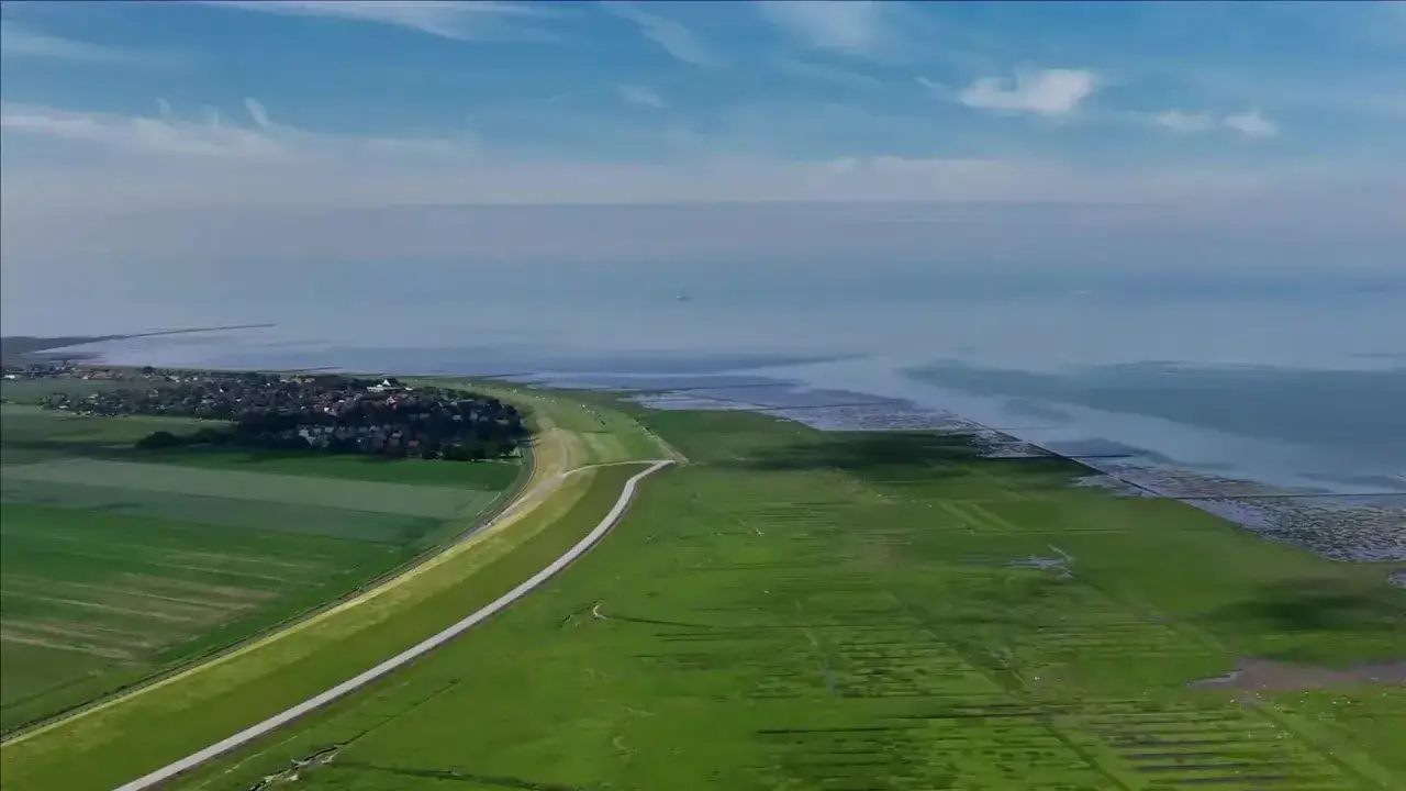 Gr&uuml;ne Felder und ein Deich sch&uuml;tzen ein Dorf in den Niederlanden vor dem steigenden Meeresspiegel.